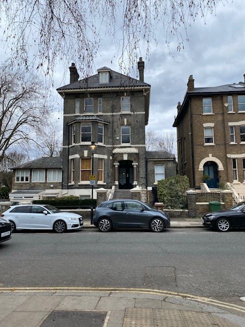 A tall, three-storey Victorian-style residential building constructed from dark grey brick with lighter stone accents around the windows and on the corners. The building features a steep, dark tiled roof with a small dormer window and two chimney stacks. Its front entrance is flanked by a small set of steps leading up to a black door, with a white lamppost situated on the pavement nearby. In front of the house, there is a narrow pavement and a street with three parked cars: a silver hatchback, a dark grey compact car, and a black sedan, positioned close to the curb. Adjacent to the right of the main building, there's a smaller structure connected via a low brick wall, partially obscured by foliage. The scene is set under a cloudy sky, with leafless trees visible above, indicating a late autumn or winter setting. This image, associated with private rubbish clearance services, captures the property in a typical urban street environment, emphasizing the residential character and local street scene.