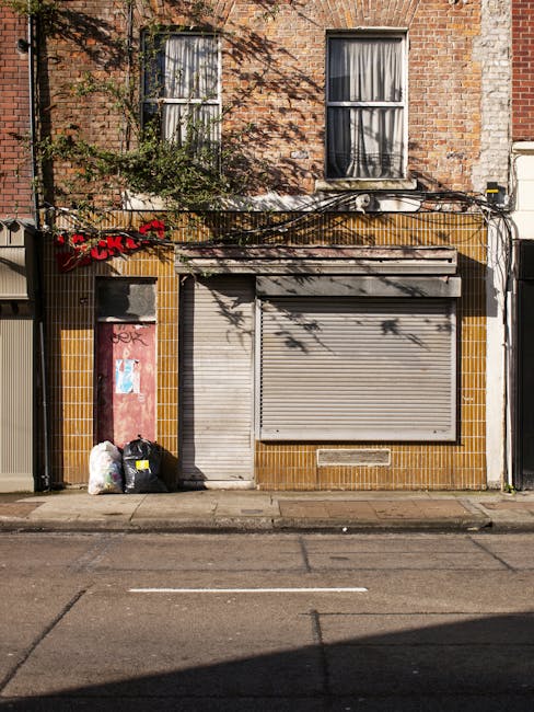 The image depicts a small, closed commercial storefront with a grey metal roller shutter covering the entrance, set within a building that has a brown tiled facade with yellow accents around the bottom section. To the left side of the storefront, there are two black and white plastic rubbish bags and a white plastic bag, placed directly on the pavement. Above, on the brick upper level of the building, there are two windows with white frames, both with metal bars across them; one window has a partially open curtain, while the other has a drawn curtain. There is some greenery, including a small tree or bush, growing close to the storefront on the left, casting shadows across the facade and sidewalk. The scene appears to be in an urban area, with a single lane of road visible in front, and the overall lighting indicates daylight conditions, possibly late morning or early afternoon. The setting suggests a location where independent waste collection services, such as those provided by companies like Rubbish Clearance Swiss Cottage, might be used for rubbish removal or on-site clearance in private or commercial premises.