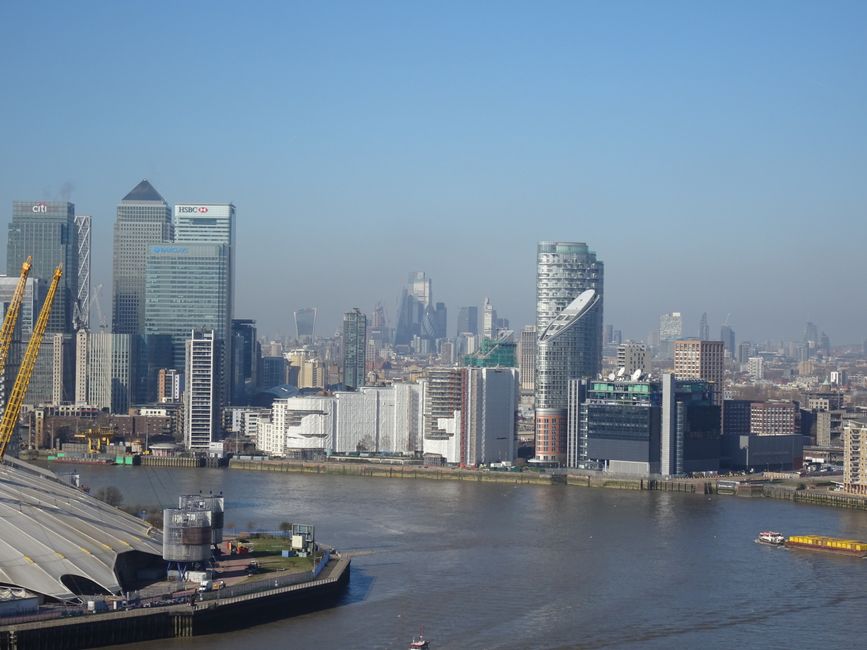 A panoramic view of the London skyline along the River Thames during daytime, featuring modern high-rise office buildings and skyscrapers with glass facades reflecting sunlight, and a few construction cranes visible on the left side. The river in the foreground has a gentle current, with a small boat navigating near the bank. The scene is set under a clear, blue sky with minimal cloud cover, and the overall atmosphere highlights urban development and skyline diversity, which relates to context of alternative waste handling and urban rubbish clearance services near central London.