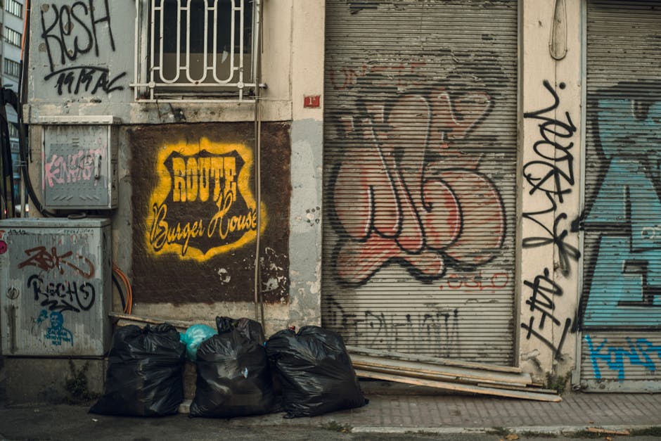 A row of five black garbage bags tightly packed and piled on a concrete pavement in front of a graffiti-covered building wall. To the left, there are electrical boxes and utility meters mounted on the wall, partially obscured by the bags. The building exterior features a dark, weathered wall with patches of peeling paint and extensive graffiti tagging in black, red, and blue, with some stylized lettering. The graffiti includes a prominent yellow sign with black lettering that reads 'BOUTIQUE Burger House,' painted directly onto the wall. To the right of the bags, there is a corrugated metal security roller shutter that is heavily graffitied in red and black, with some faded blue and black additions. A few wooden planks are placed horizontally on the ground next to the bags, leaning slightly against the shutter. The scene takes place outdoors, possibly in an urban or industrial area, highlighting an example of private rubbish awaiting collection or disposal, reflecting alternative waste management methods. The lighting appears natural, with diffused daylight casting soft shadows across the surface.