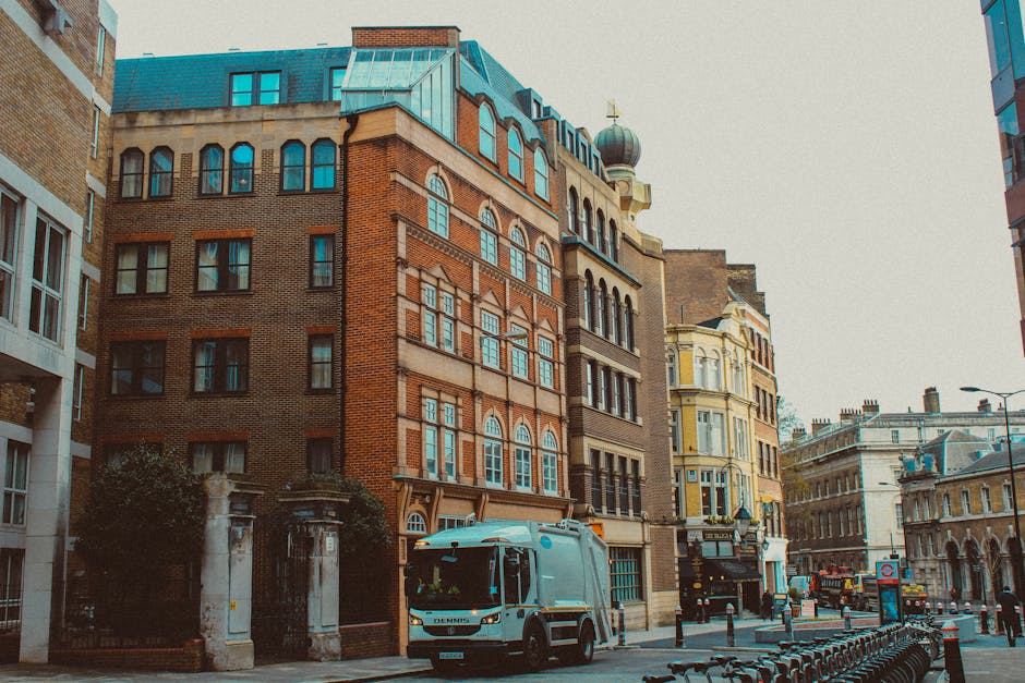 A street scene in an urban area features several multi-storey buildings with varied architectural styles, including a prominent red brick structure with arched windows and decorative stone bands. To the right, there is a cream-colored building with bay windows and intricate detailing, adjacent to more contemporary structures with large glass windows and modern facades. In the foreground, a light blue waste collection vehicle is parked on the street, partially obscuring a gated entrance with stone pillars and ornamental fences, which suggests a private property or institutional access point. The street is lined with metal bike racks and bollards, with some pedestrians and vehicles visible in the background, under an overcast sky that provides diffuse lighting, emphasizing the textures of the brickwork, stone, and modern glazing. This scene implicitly relates to local waste management services, with the presence of the waste collection truck indicating activities related to rubbish removal and private disposal handling in the area.